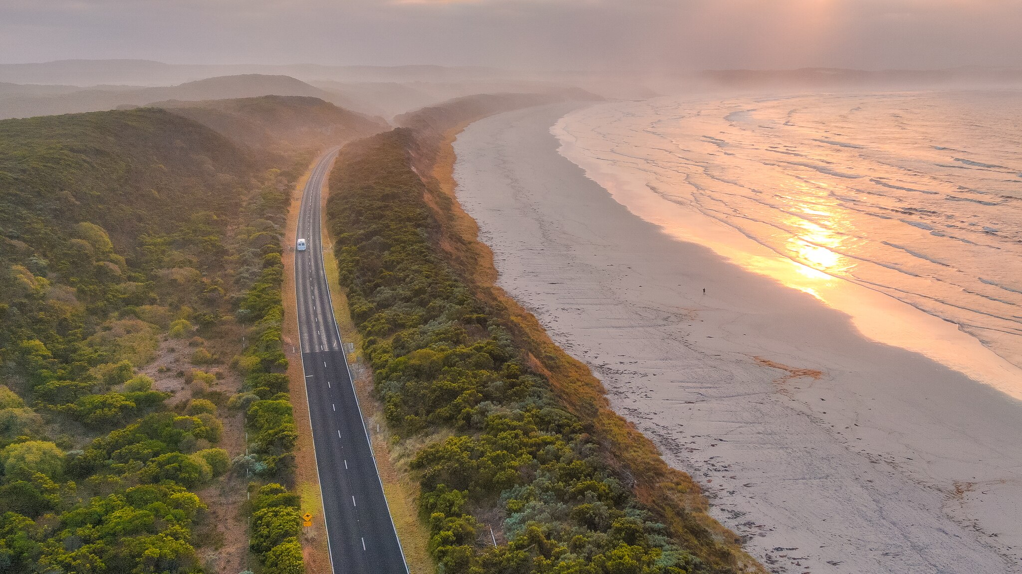 A rising sun casts skies of pink and yellow over a coastline bordered by lush green vegetation.
