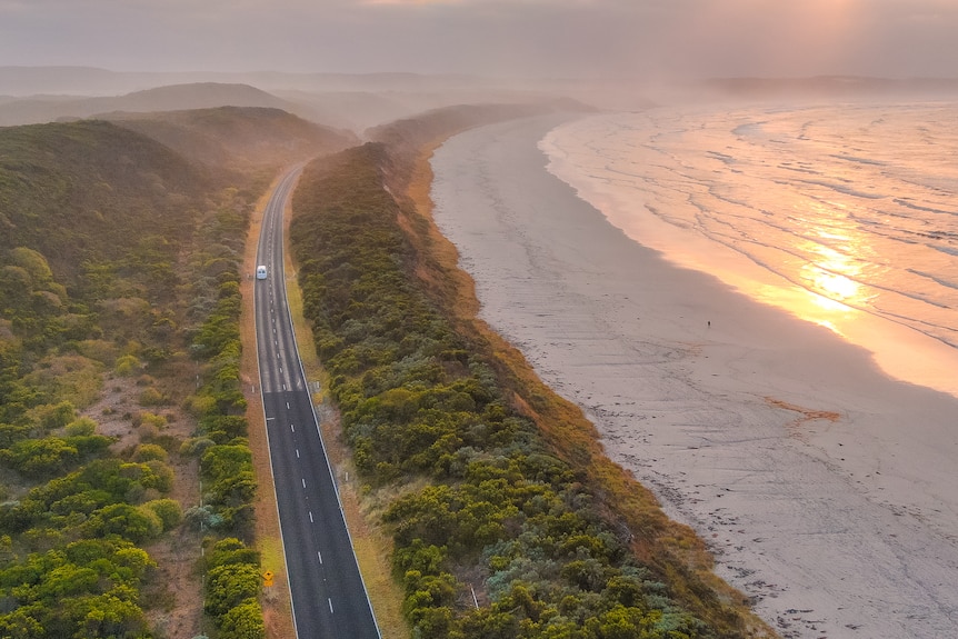 A rising sun casts skies of pink and yellow over a coastline bordered by lush green vegetation.