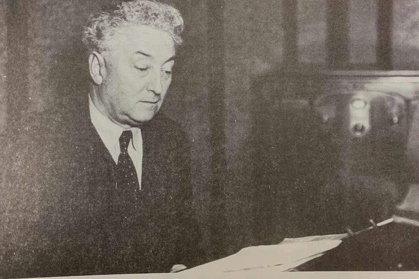 Black and white photo of Prime Minister Lyons in a suit in a studio, reading off a piece of paper