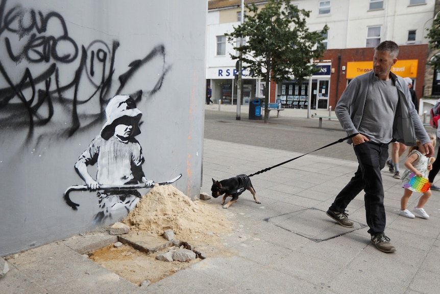 A man and his dog walk past an artwork created by Banksy on a building corner in Lowestoft, England