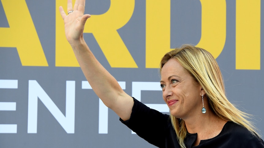 A woman with blonde hair wearing a black dress waves in front of a grey background with yellow and white writing. 