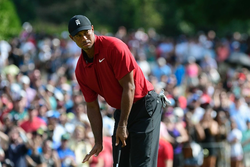 A man wearing a red shirt motions with his right hand while bent over, holding a golf club in his left