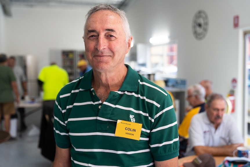 Man in a green shirt standing in front of tables of men having smoko in a men's shed, smiling kindly