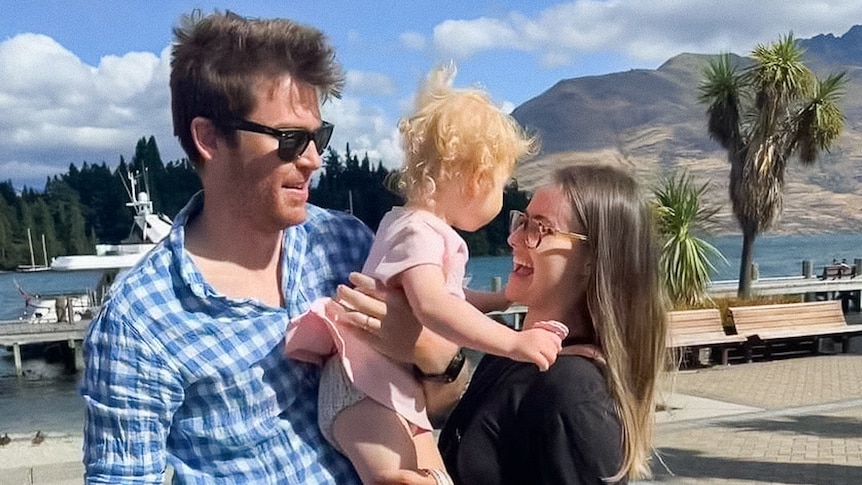 A woman with long brown hair smiles at her toddler daughter behind held by her husband beside her.