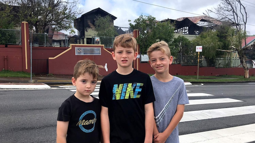 Three primary school-aged boys stand outside their school with fire-damaged buildings in background.