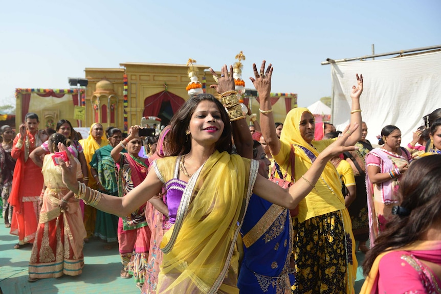 A group of colourfully dressed hijras dance, arms raised, down the street in a religious procession.