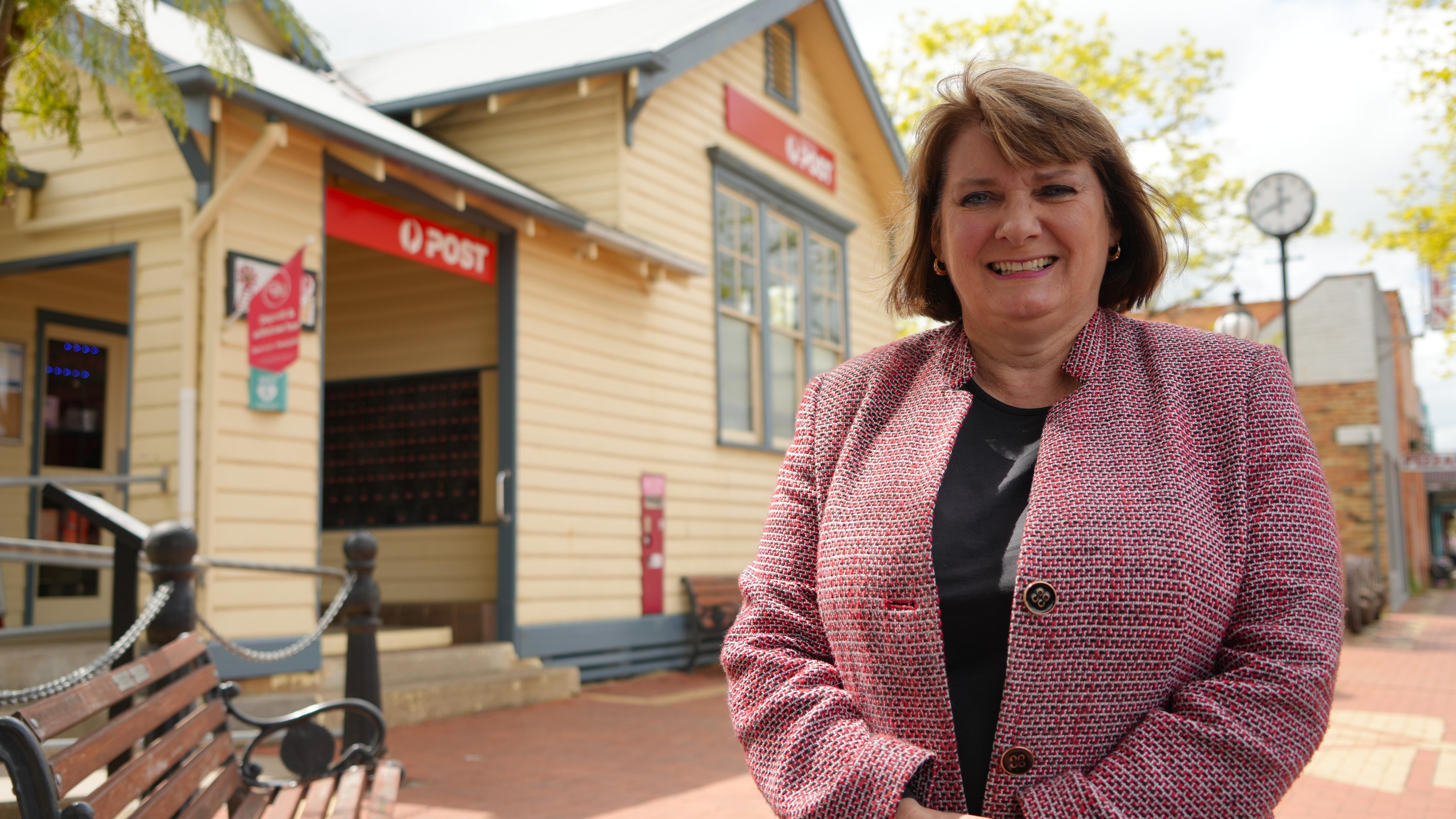 A smiling woman with a short brown bob standing in front of a town post office.