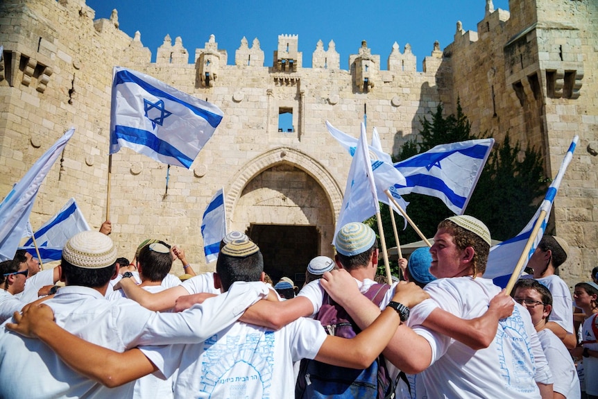 A large crowd of people, including many waving Israeli flags, outside a large building.