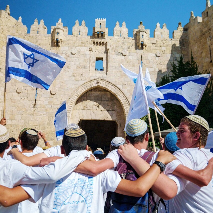 A large crowd of people, including many waving Israeli flags, outside a large building.