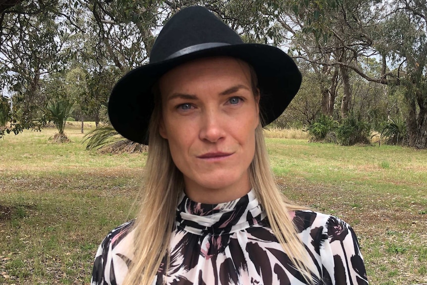 A head and shoulders shot of a woman with blonde hair and a black hat standing in front of bushland.