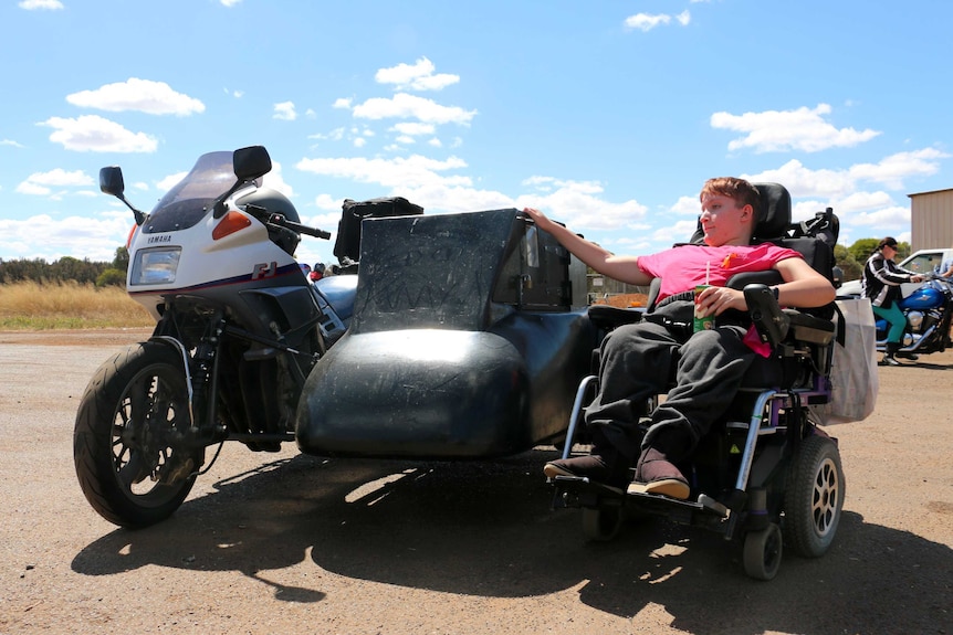 Katanning teenager Shanniah Barker sits in her wheelchair next to her father's motorbike, holding onto its sidecar.