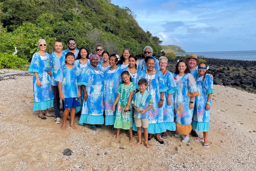 A large family group dressed in blue island dresses and shirt on a beach with rocks and mountain in background