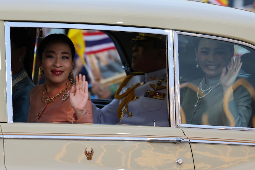 Two women wearing ornate jewellery and pastel colours wave through a car window