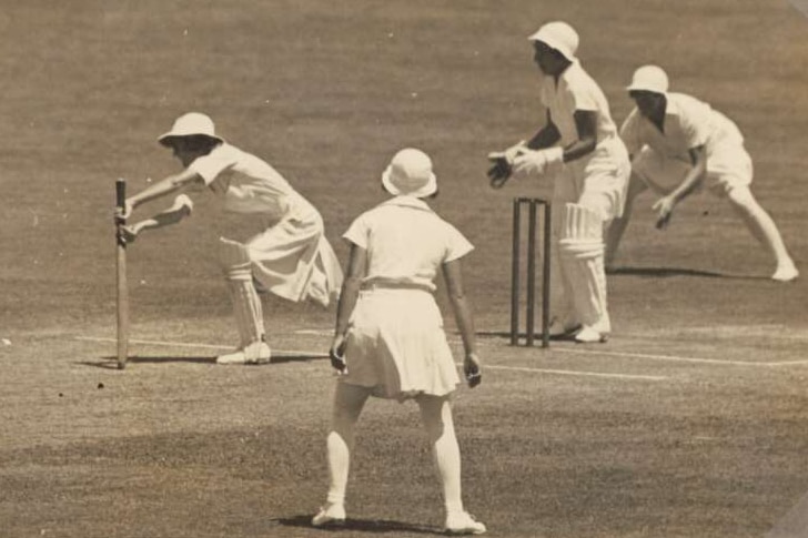 A black and white photo of cricketers out on a pitch, one is batting and the others are crowded around ready for a catch.
