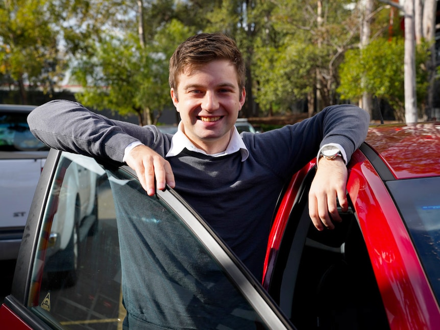 A man stands next to his car with one arm resting on the open door and one resting on the roof.