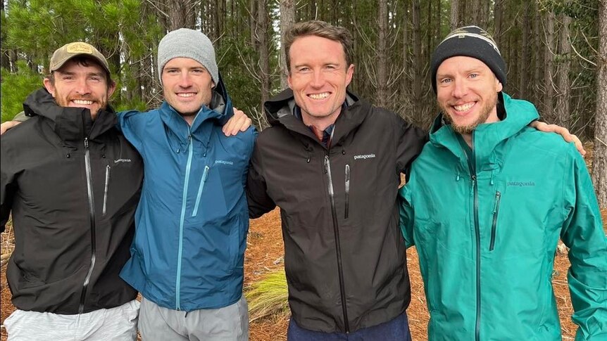 Four men in shorts and wind breakers arm in arm in a forest