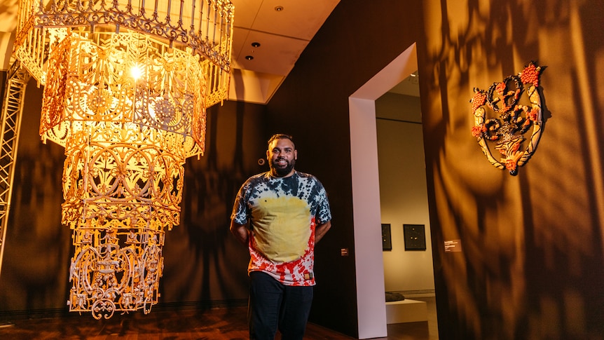 Warrang man with short dark hair and beard smiles wearing a tie-dyed Aboriginal flag print shirt beside large yellow chandelier