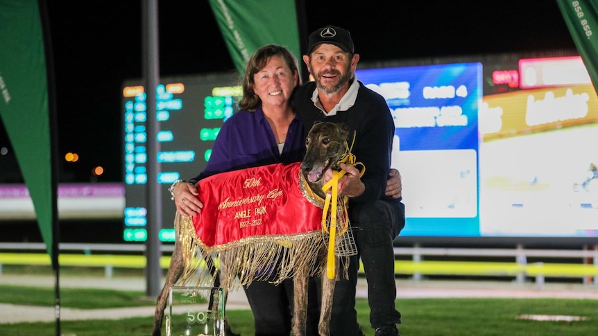 A woman and man standing with their hands resting on a greyhound with a trophy in front