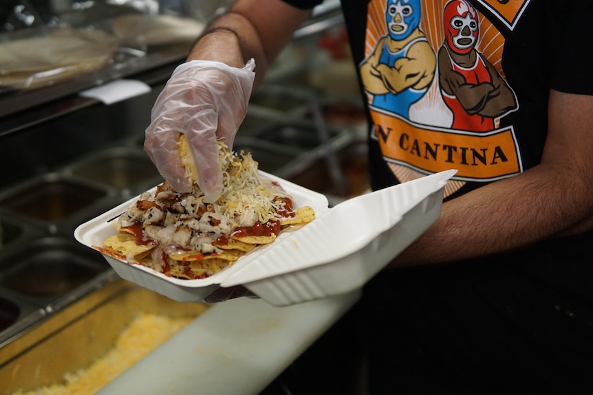 A gloved hand puts cheese on a box of nachos.