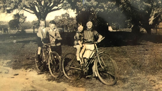 Sepia coloured old photo of two young children on bikes, with two older children standing next to them supporting them.