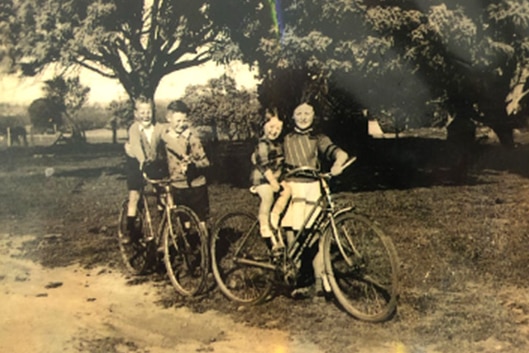 Sepia coloured old photo of two young children on bikes, supported by two older children standing beside them.
