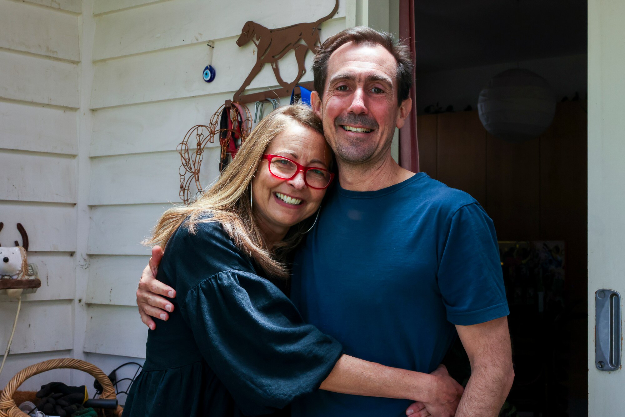 A man and a woman stand with their arms around each other, at the front door of their home.