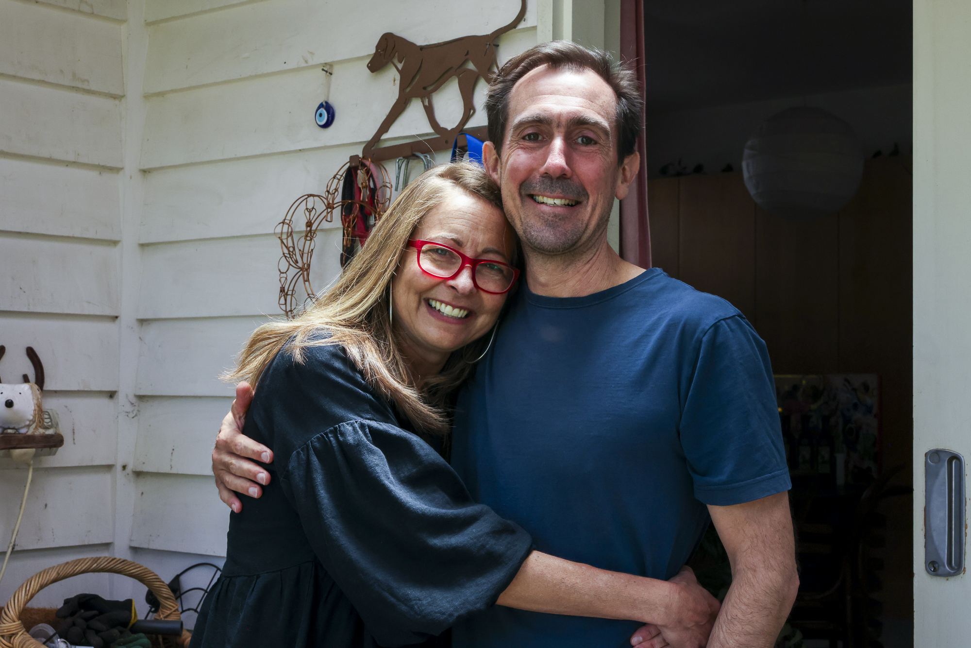 A man and a woman stand with their arms around each other, at the front door of their home.