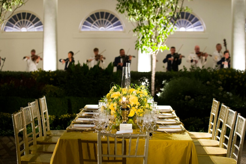 A row of violinists playing behind an elegantly set table covered in yellow flowers