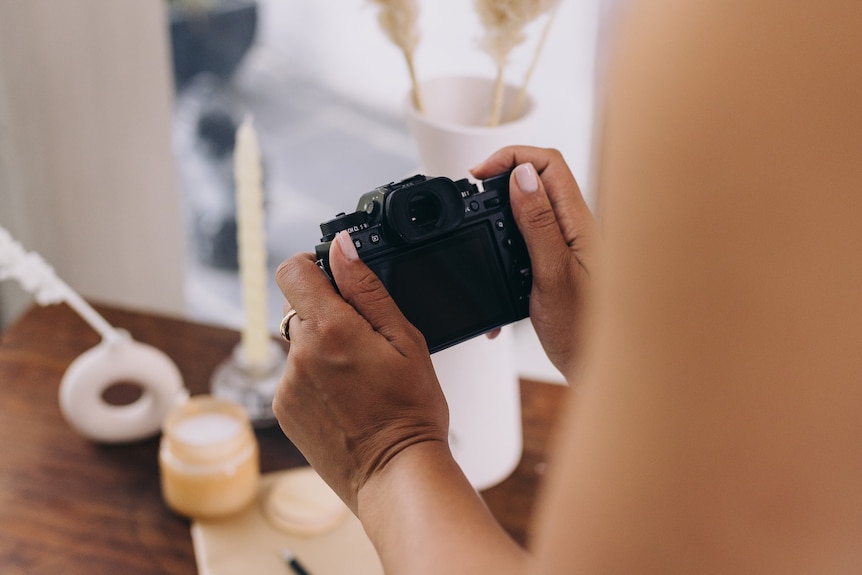 Woman with dark skin takes photos of a candle and flowers on a work table. 