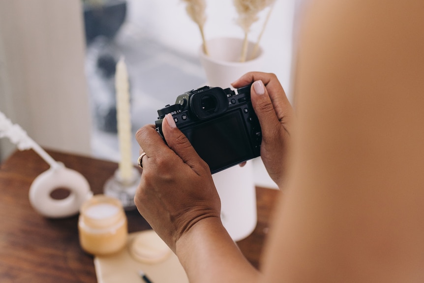 A dark-skinned woman is photographing candles and flowers on a workbench. 