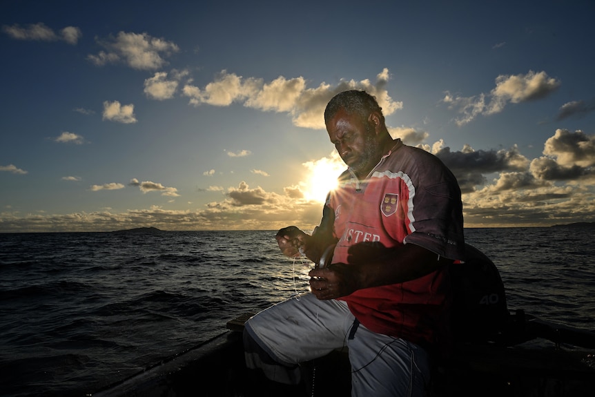A man sits in a fishing boat at sea with the sun behind him as a he prepares a line and hook