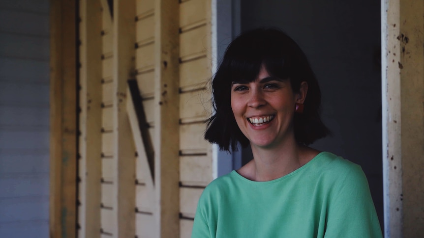 A woman with short brow hair in a green top, smiling in front of her house
