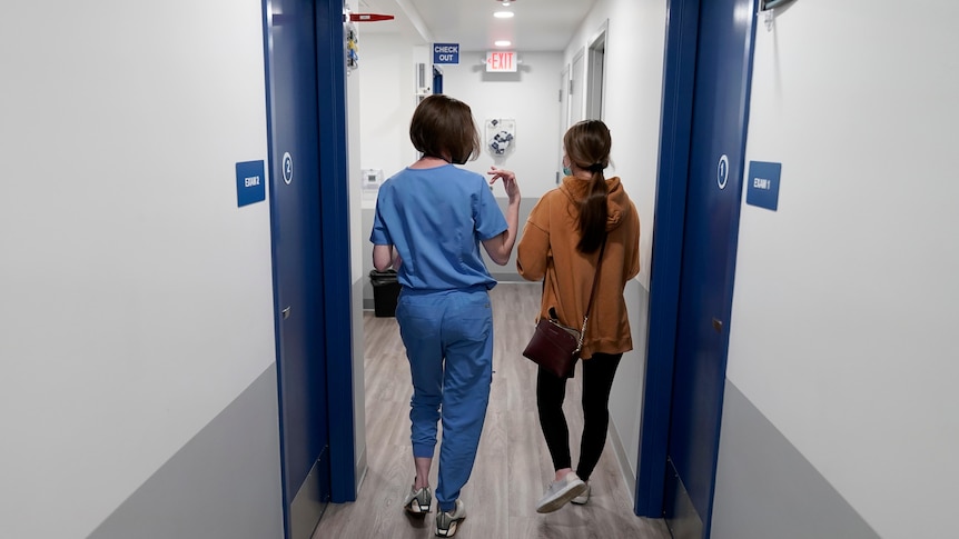 A woman in scrubs and another woman walk down a clinic hallway. 