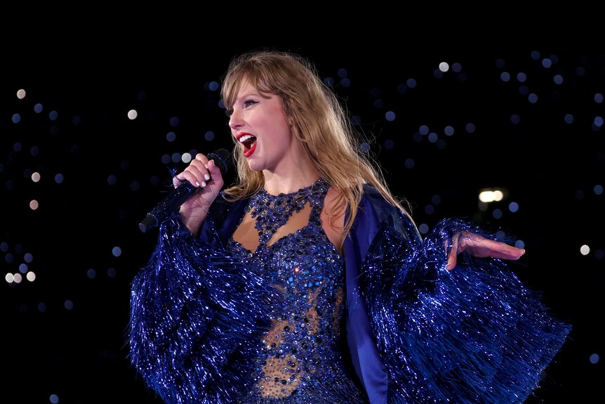 Taylor Swift sings into a microphone as she fans her left hand out beside her while dressed in a blue bodysuit and tinsel jacket