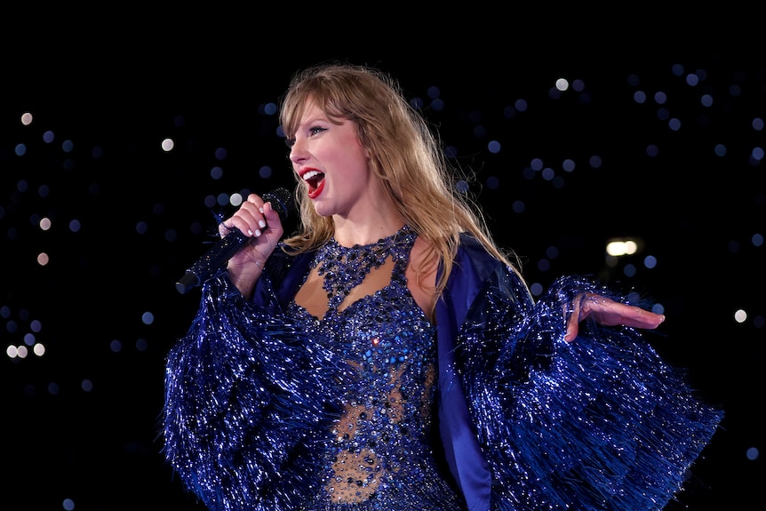 Taylor Swift sings into a microphone as she fans her left hand out beside her while dressed in a blue bodysuit and tinsel jacket