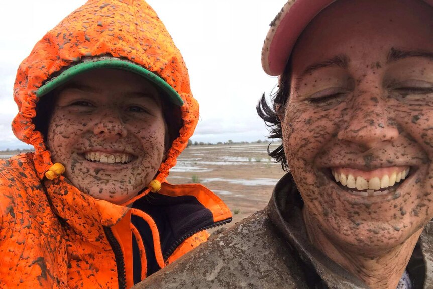 Two women splattered head to toe in mud smile widely at the camera, woman on right has eyes closed