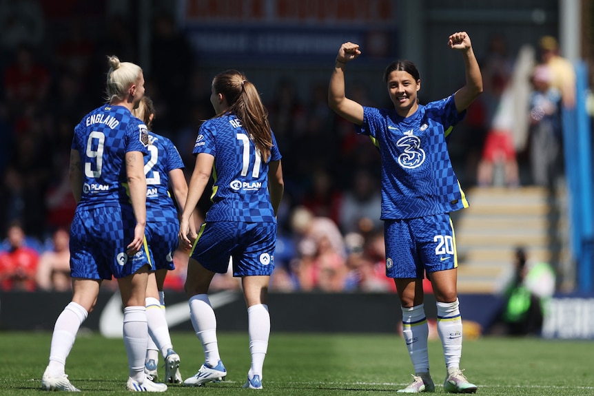 Sam Kerr holds her hands in the air as she smiles on the field.