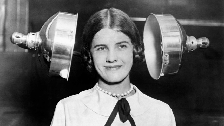 A vintage black and white photo of a woman having her hair dried by two large metallic heating lamps.