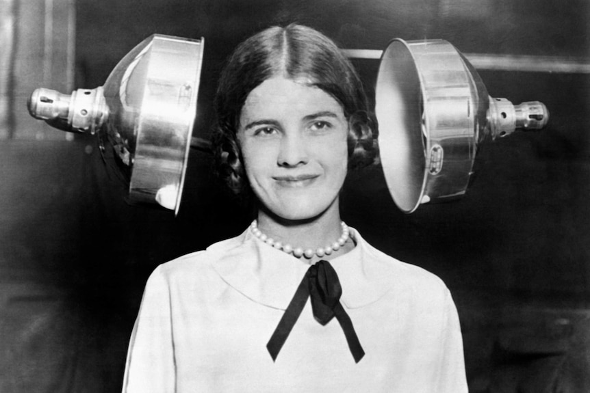 A vintage black and white photo of a woman having her hair dried by two large metallic heating lamps.