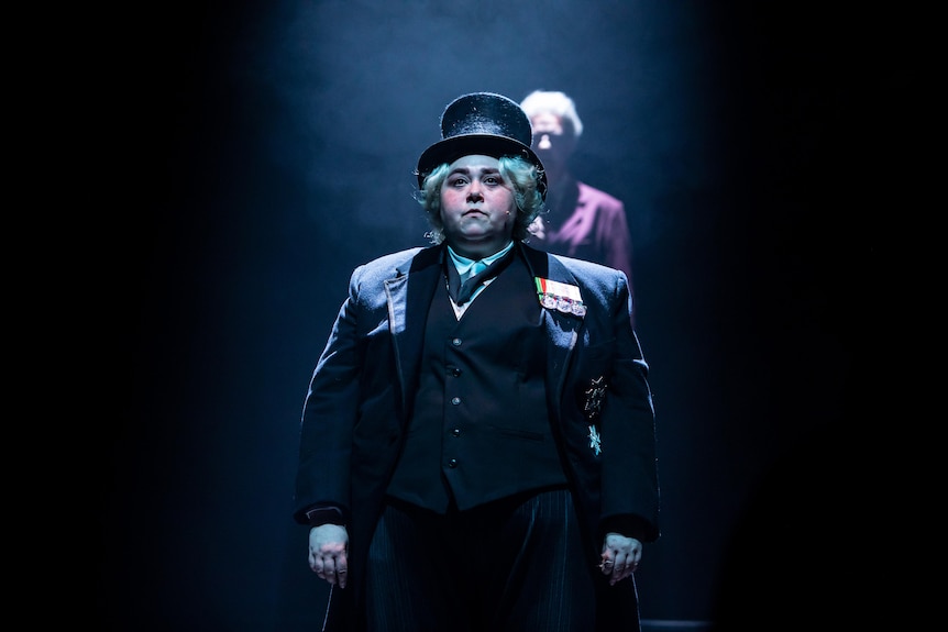 Octavia Barron-Martin onstage as Sir John Kerr, dressed in a black three-piece suit and top hat and looking sombre