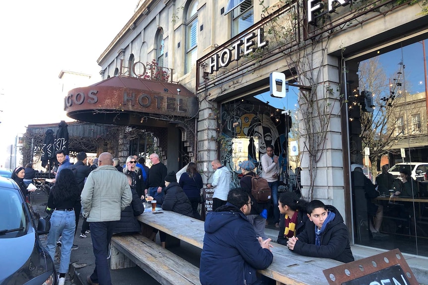 People sit and stand outside a pub hotel on a bright day drinking beer and eating food.