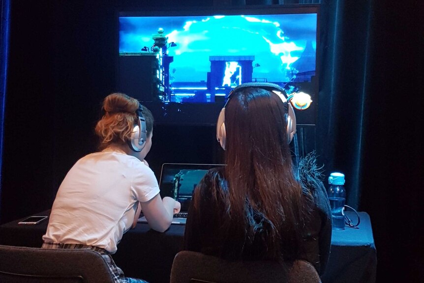 Two women with headphones on play a game at a computer