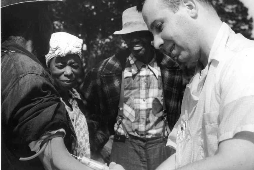 A man injects another man in the arm, as two other people look on.