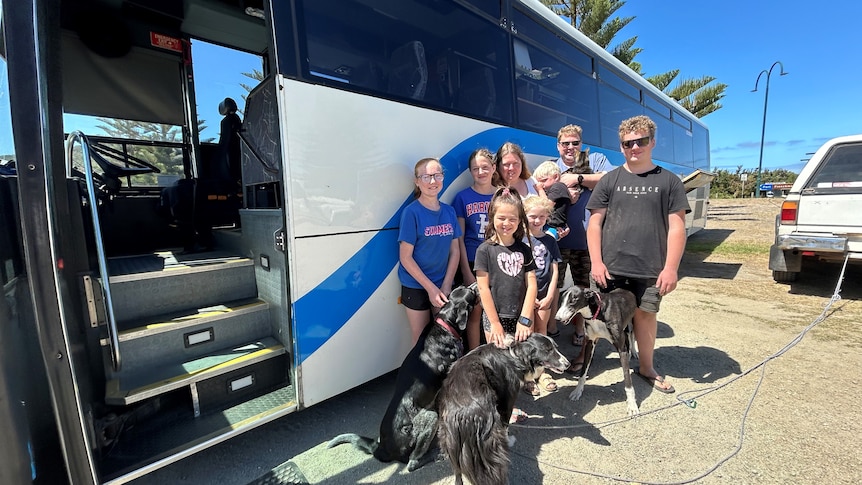 A family of 8 stands in front of a large blue and white bus with three dogs and one cat. 