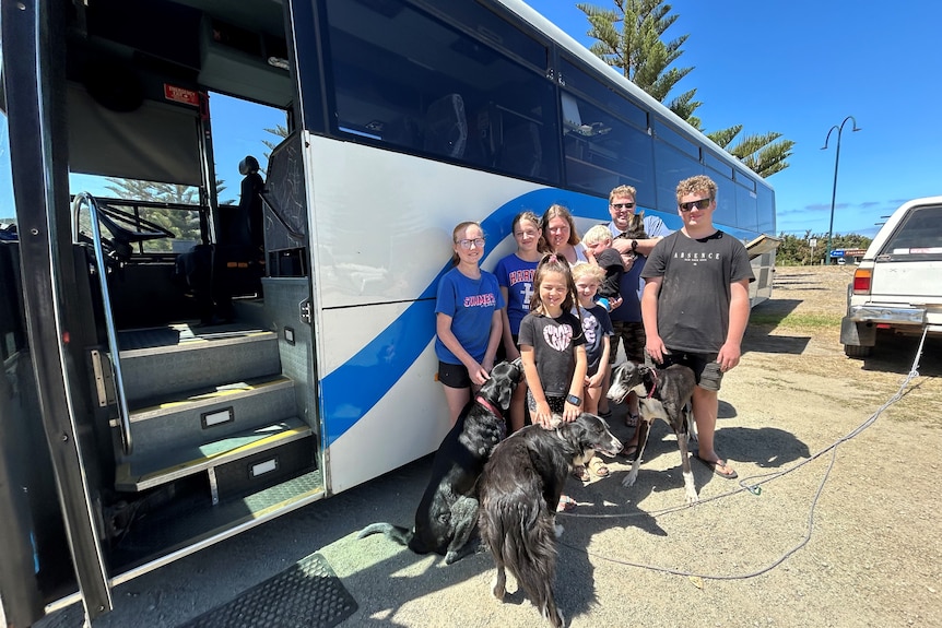 A family of 8 stands in front of a large blue and white bus with three dogs and one cat. 