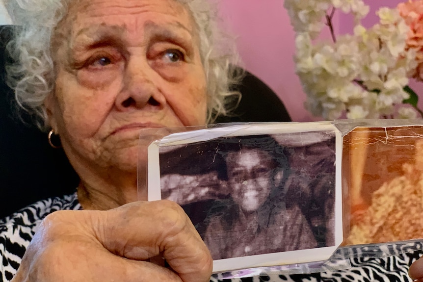 An elderly lady holds up two faded photos of a small, smiling boy