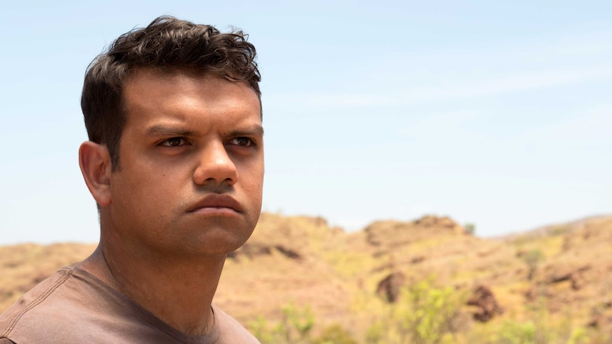 A young Aboriginal man stares off into the distance, arid nature in the background