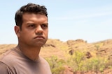 A young Aboriginal man stares off into the distance, arid nature in the background