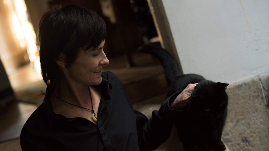 A woman with short black hair is sitting on grey carpet and patting a black cat