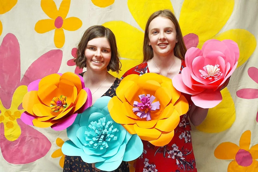 Two girls holding large paper flowers in front of a background painted with yellow flowers.